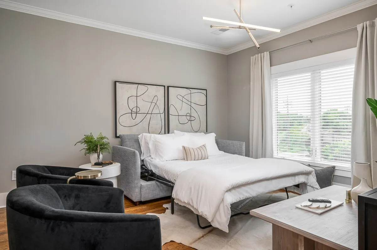 The image showcases a bright and stylish interior bedroom, featuring a plush bed with soft linens, modern artwork on the walls, two inviting chairs with a side table, and a large window flooding the space with natural light. The setting is perfect for relaxing, reading, or enjoying quiet time, with a cozy ambiance enhanced by thoughtful decor.