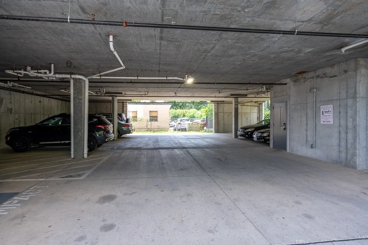 An interior shot of the parking area or garage featuring parked cars, a visible garage entrance, and concrete walls. The mood is practical and utilitarian, emphasizing the convenience of on-site parking and ease of loading or unloading luggage.