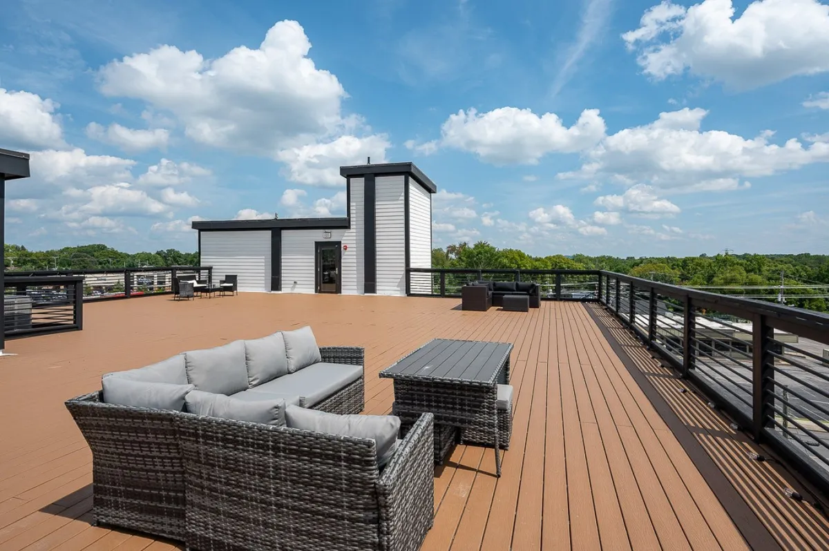 A spacious rooftop deck with comfortable outdoor seating overlooking the city skyline and surrounded by lush greenery. The scene features people relaxing, socializing, and enjoying the panoramic views, with the open sky creating a vibrant, inviting atmosphere.