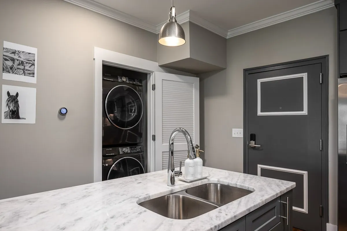 A contemporary utility area featuring a marble countertop, stainless steel sink, modern appliances, and wall art. The setup is ideal for both light laundry and household tasks, reflecting convenience and style.