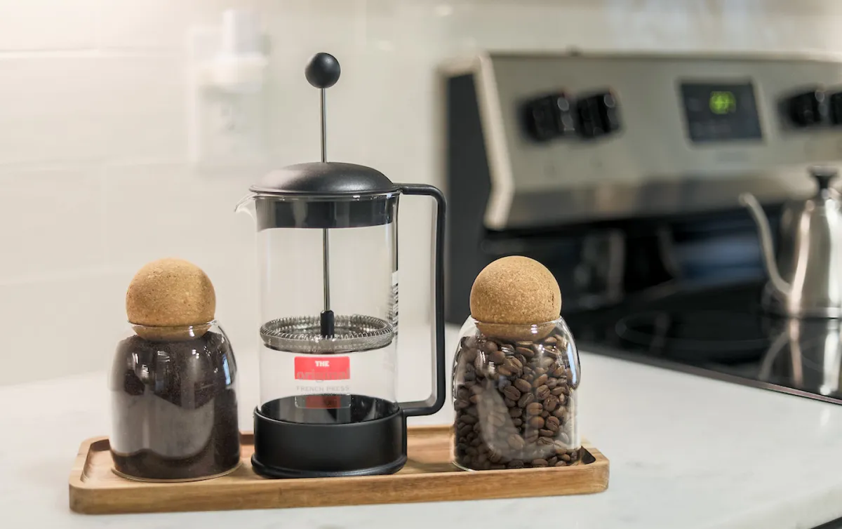 A close-up of a French press coffee maker, glass jars with cork lids, and a wooden tray, set in a cozy, sunlit kitchen corner. The setup evokes a sense of leisurely morning routines and social coffee moments.