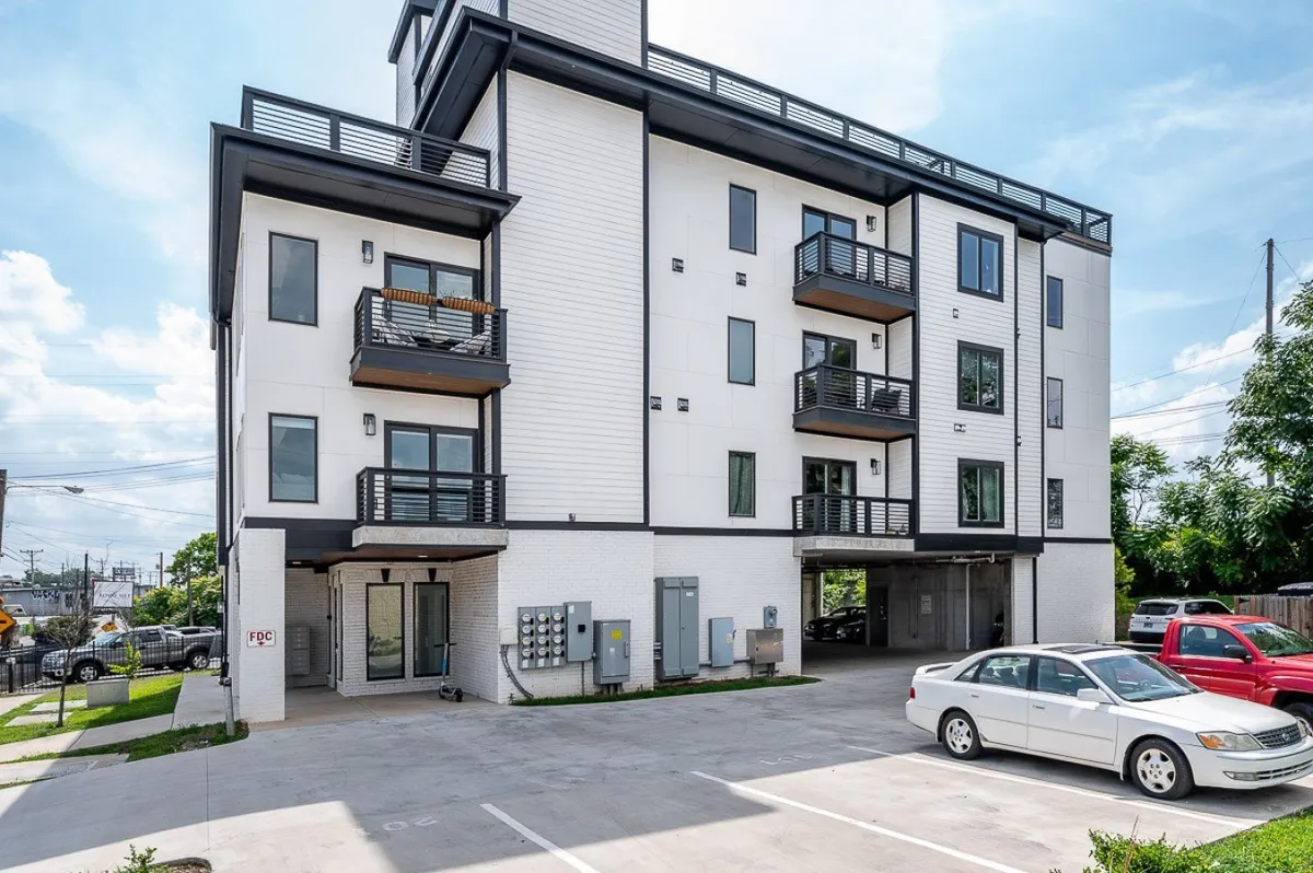 This vibrant exterior image captures the townhouse's main entrance, visible balconies, and the parking area where vehicles are parked. The scene conveys easy access to the property, privacy for arrivals, and the convenience of on-site parking.
