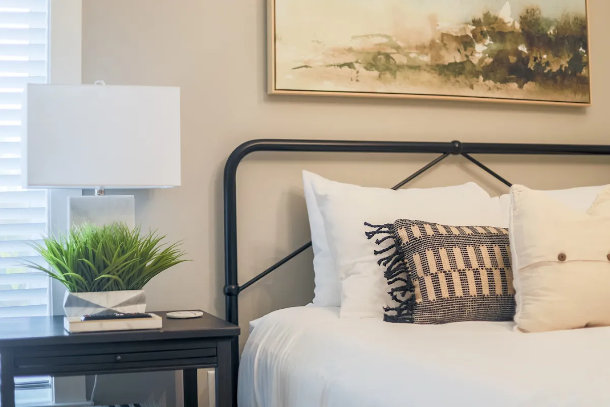 This cozy bedroom features a bed with decorative pillows, a table lamp, artwork above the bed, and a nightstand with a book and plant. The space feels inviting and restful, ideal for both solo downtime and couples.