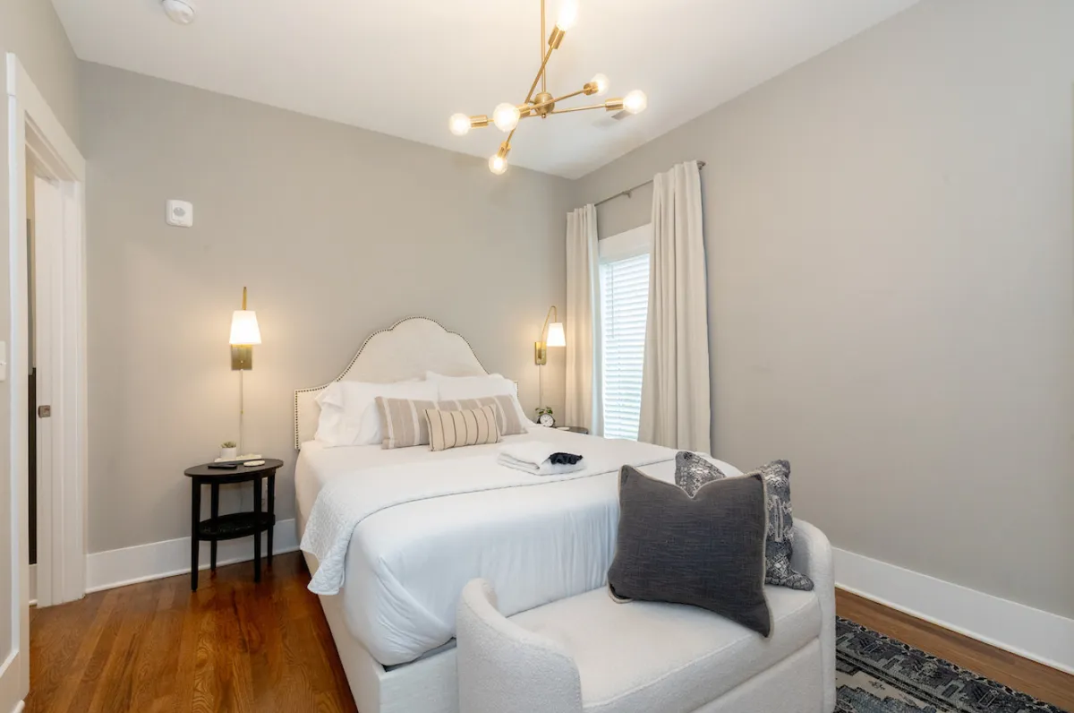 This image depicts a tranquil bedroom with a bed adorned with decorative pillows, a modern light fixture, and a textured rug. Soft natural light filters in, creating an inviting atmosphere ideal for rest and relaxation.