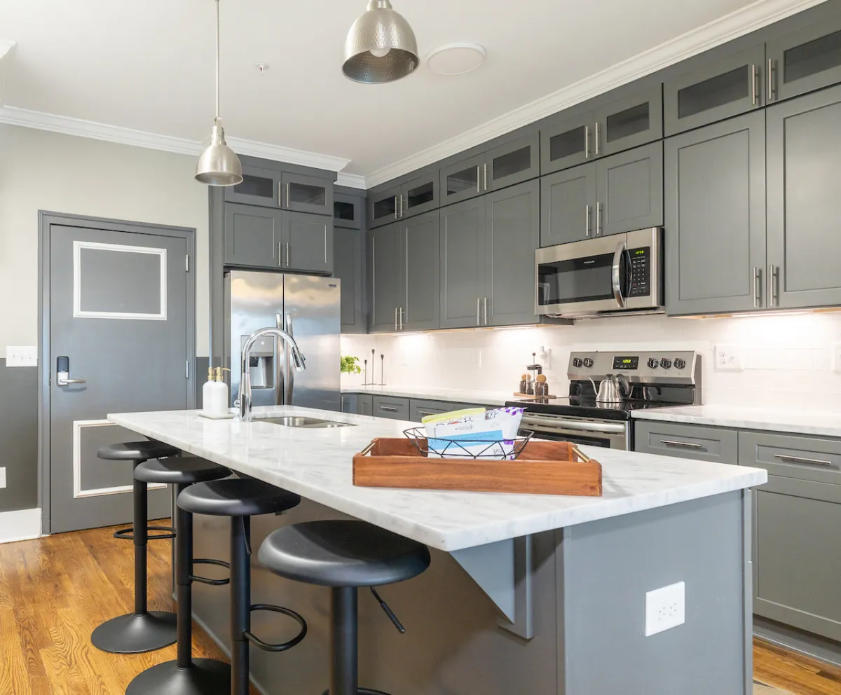 A modern kitchen scene with a central island surrounded by bar stools, stainless steel appliances, sleek cabinetry, and stylish pendant lighting. The layout is open and flows directly into the living area, emphasizing the functional and social aspects of the space.