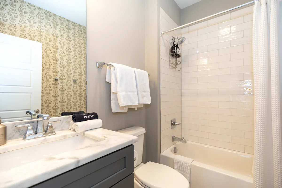 A modern bathroom is highlighted by a marble countertop, a sleek faucet, decorative wallpaper, and towels neatly arranged for guests. The space feels calm, clean, and designed for self-care.